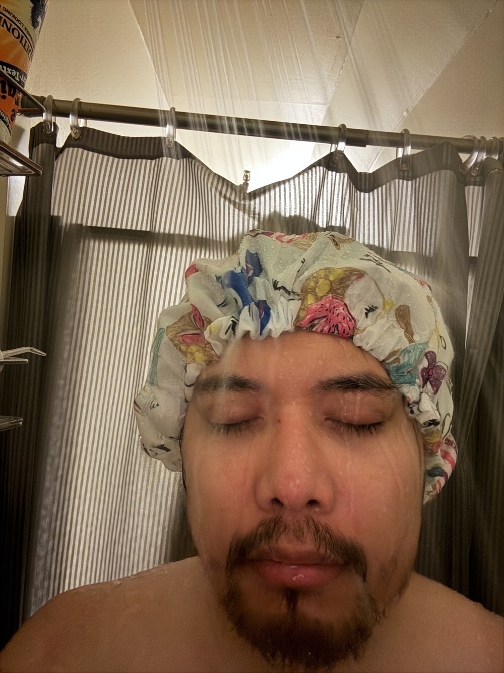 A smiling person wearing a floral shower cap is taking a shower, holding up a peace sign with their right hand.