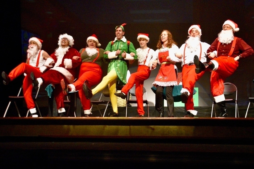 A group of people on stage wearing festive Christmas costumes including Santa Claus outfits and elf attire, performing a kick line.