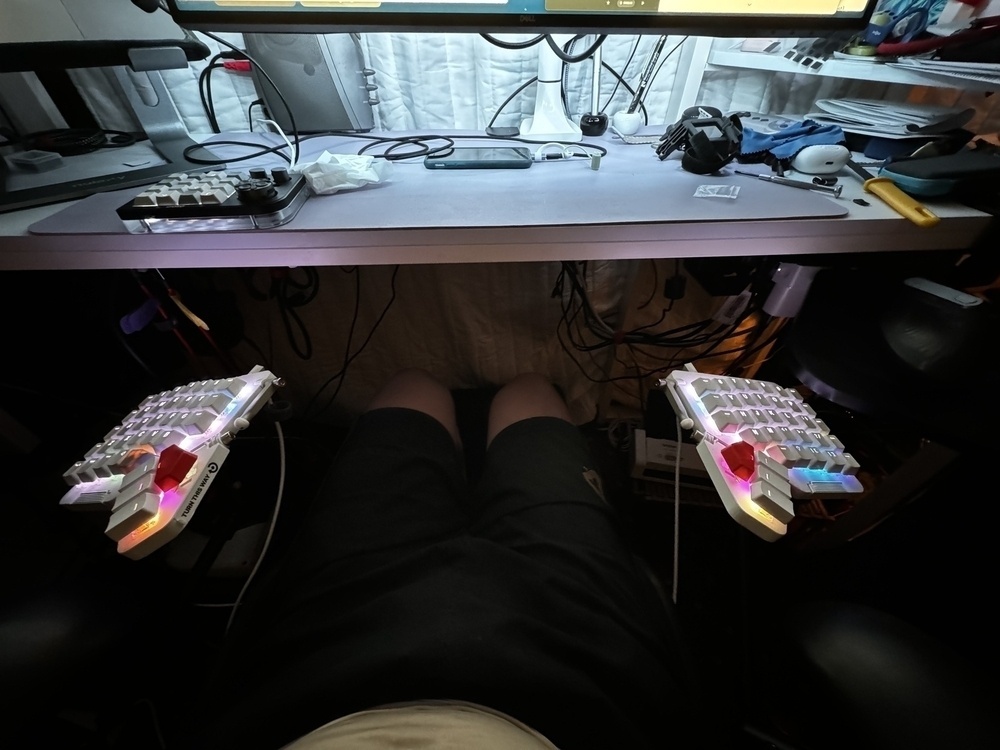 The image shows a seated person in front of a desk setup. The person's legs are visible in the foreground. The desk above has various items, including a monitor, a game controller, some cables, and tools. The focus is on two custom mechanical keyboards, each with RGB lighting and attached to adjustable mounts on either side of the person’s thighs, allowing ergonomic positioning for typing. The overall environment is a mix of tech equipment and workspace items.