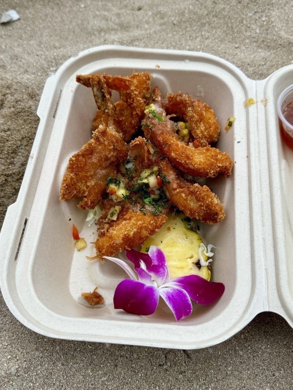 Fried shrimp garnished with herbs and a purple flower, served in a take-out container, on a sandy surface.