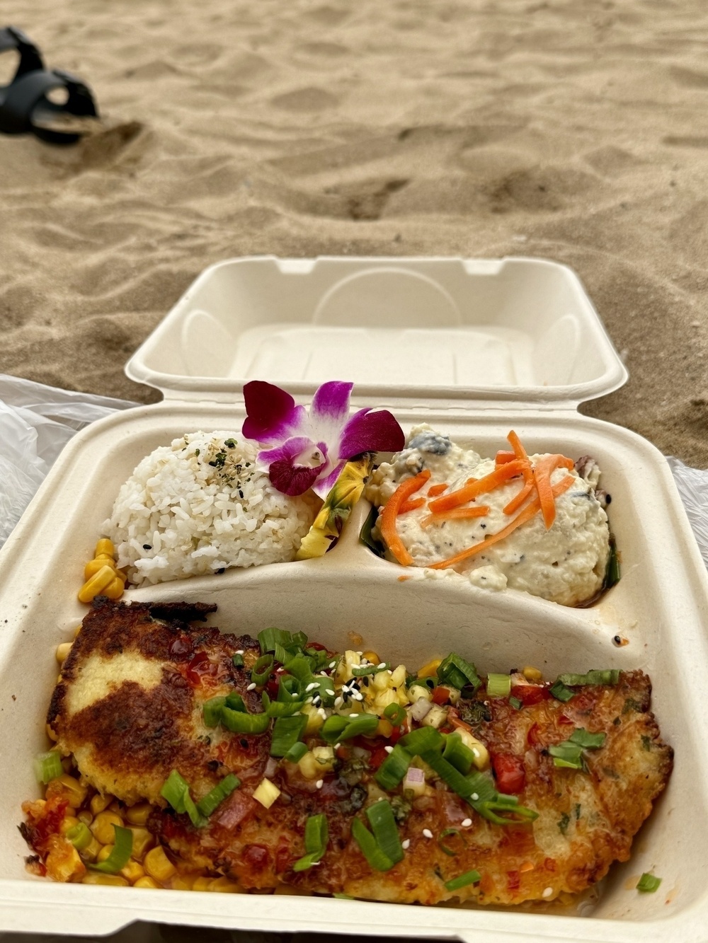 A takeout meal with rice, a flower garnish, and two entrees rests open on sandy ground.