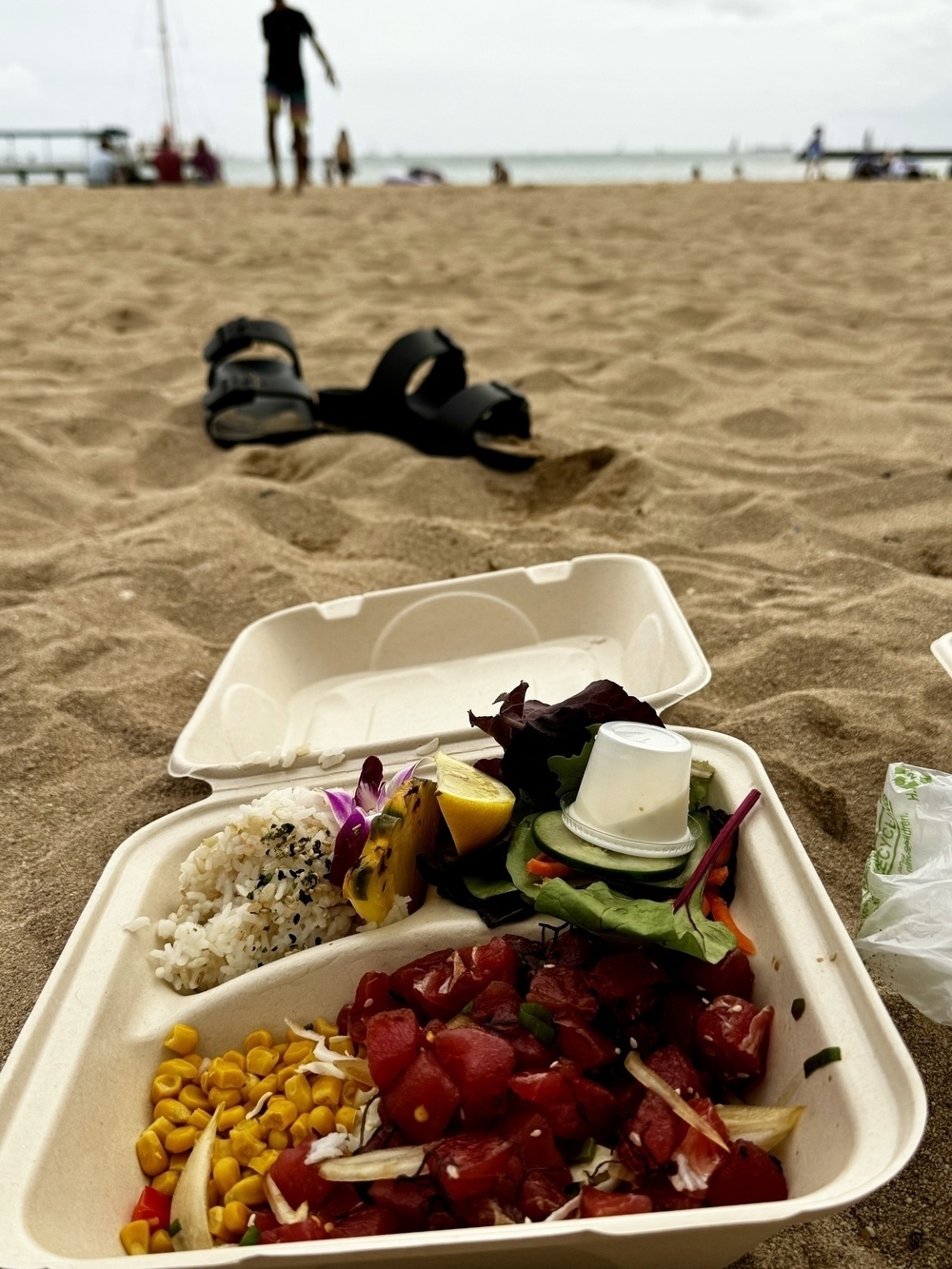 An open takeout box filled with food rests on the sand, with beachgoers and the ocean in the background. A pair of sandals lies nearby.