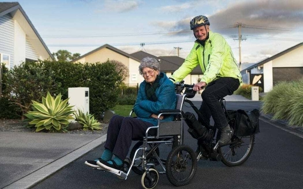 Wheelchair attached to front of bike; woman in chair, man on bike. 