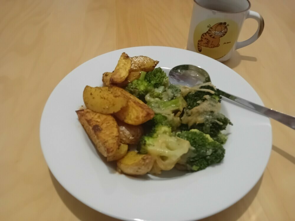 Ziefer weißer Teller mit links einer Portion Kartoffelwedges, rechts einer Portion überbackenem Brokkoli, am Rand ein Esslöffel, oberhalb des Tellers steht eine Tasse mit einem grinsenden Garfield drauf. 