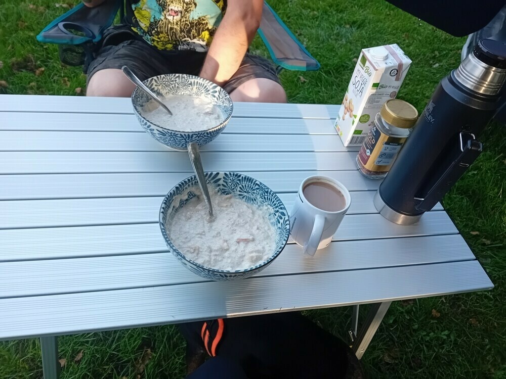 Alu-Campingtisch, darauf stehen zwei Speiseschalen mit Müsli, eine Tasse mit Kaffe, eine schwarze Thermoskanne, ein Glas mit Krümelkaffee und eine Packung Sojamilch. Dahinter sitzt der Mann in einem Campingstuhl, von dem sieht man aber nur Beine, den linken Arm und einen Teil des Rumpfs. 