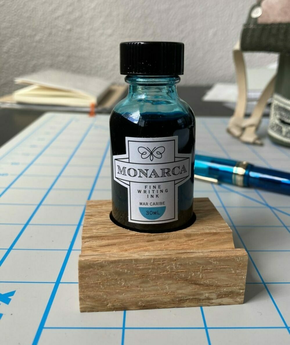 bottle of Monarca Mar Caribe fountain pen ink in the little wooden stand that came with the ink. on a messy table, with notebooks and other pens in the background.