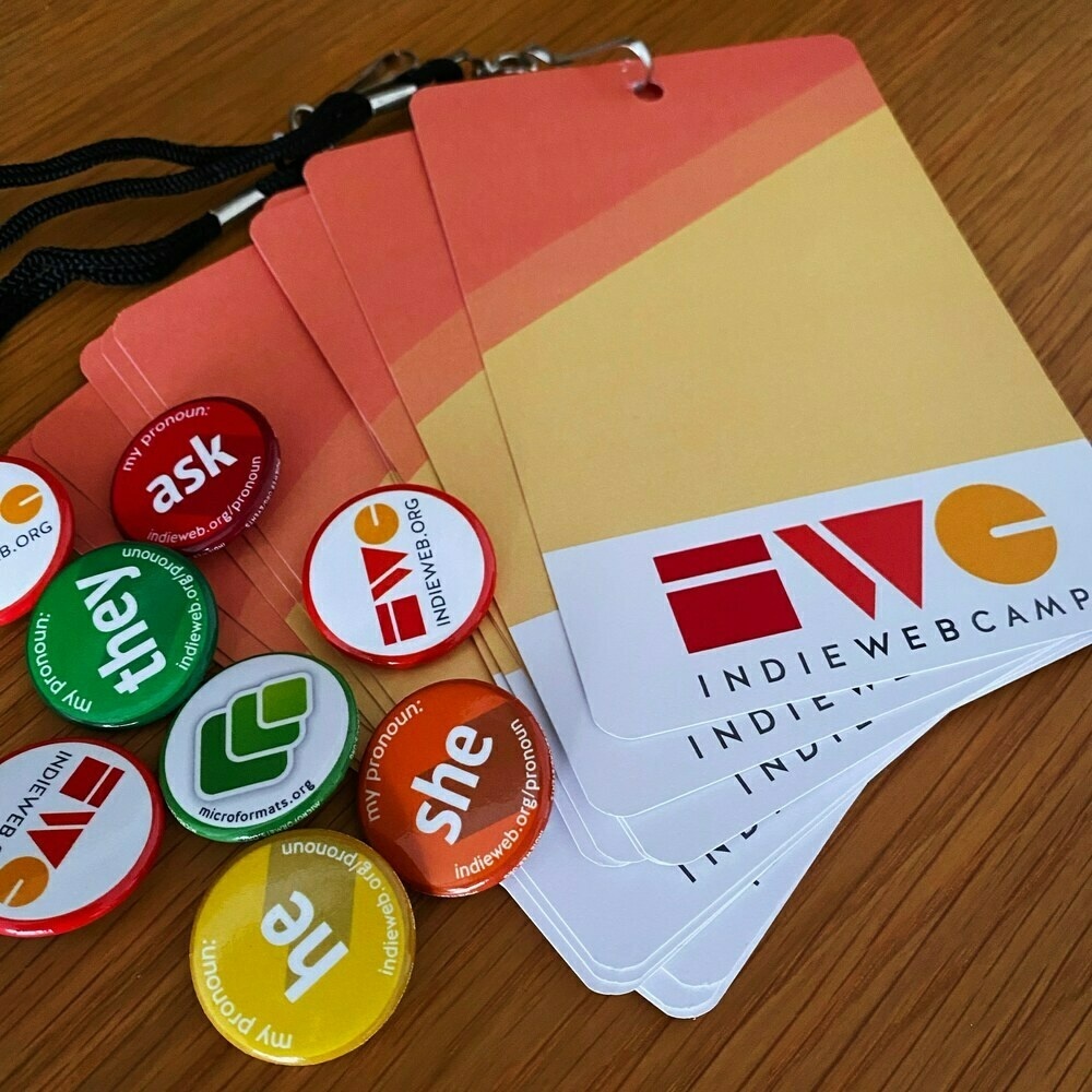 Small coloured pin badges for different pronouns sit on a stack of blank lanyards with the IndieWebCamp logo on them.