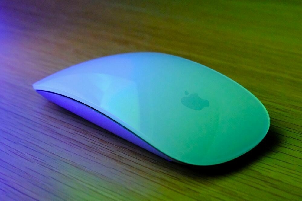 An Apple Magic Mouse on a wooden desk, highlighted by the glow of nearby green and purple lights.