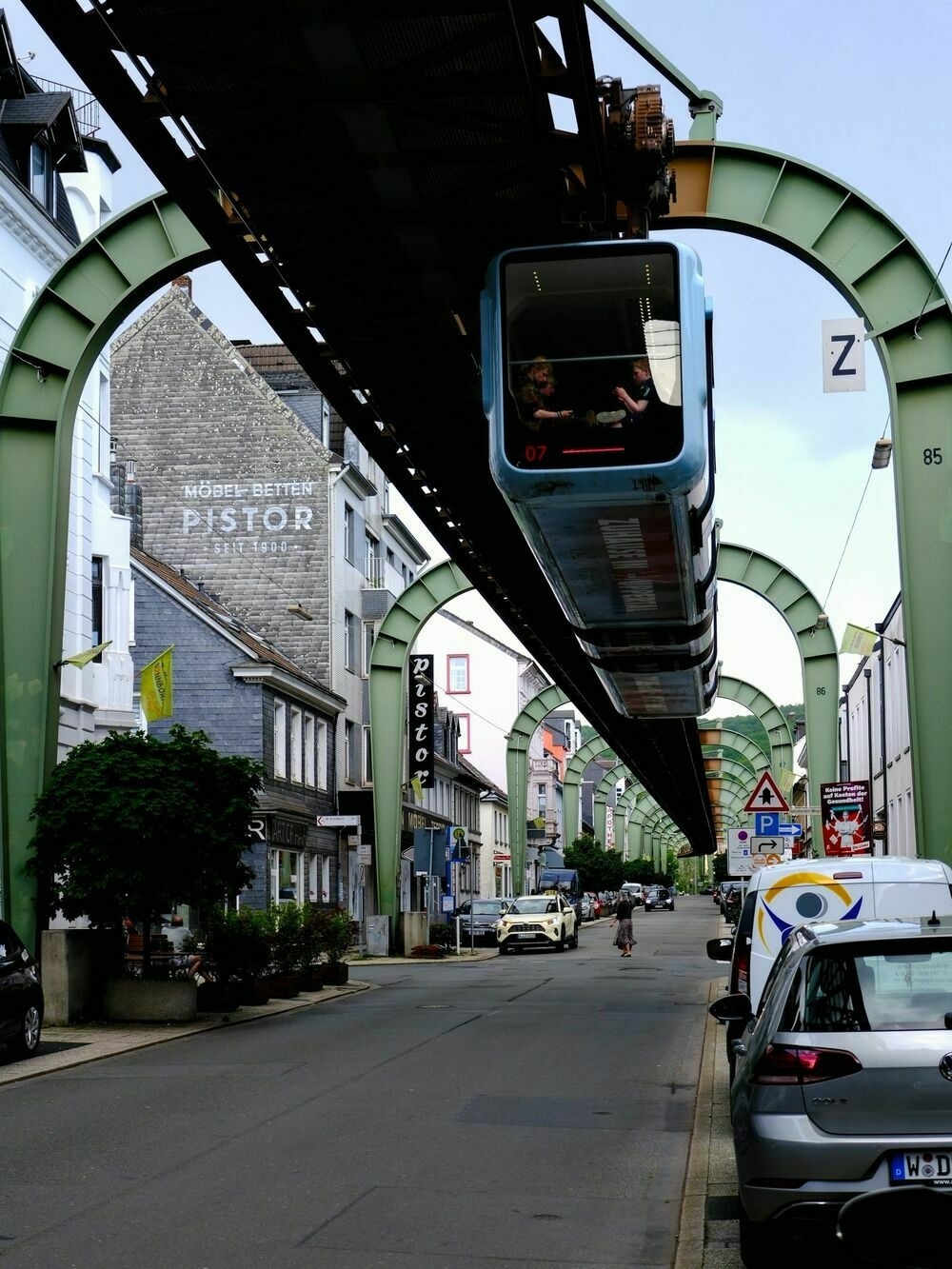 A suspended railway with a blue train carriage suspended from it passes over a quiet street with cars parked along one side.