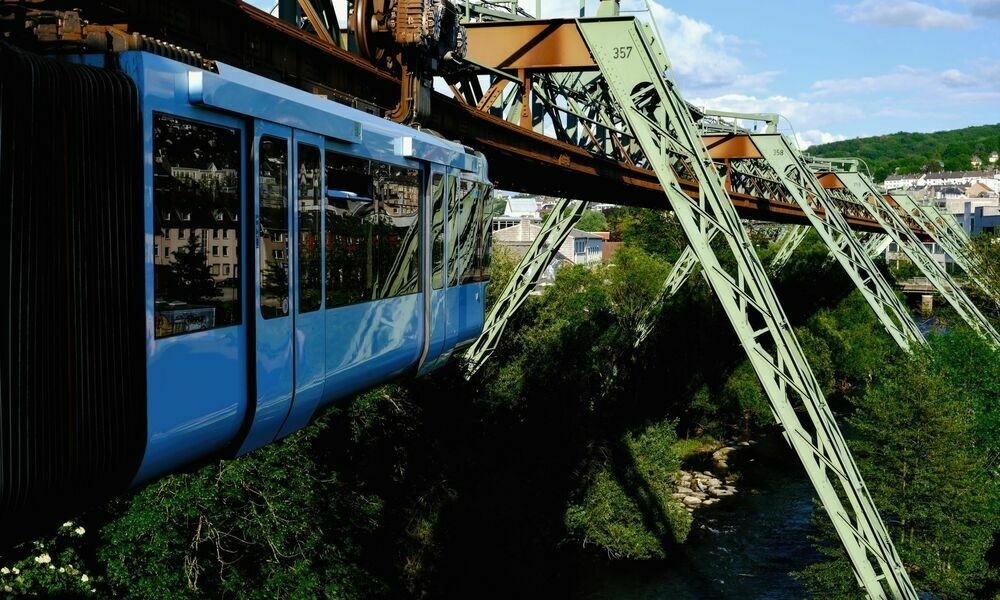 Close up of a train carriage as it pulls out of a station. The winding track ahead can be seen with the railway suspended over a river by light green metal supports.