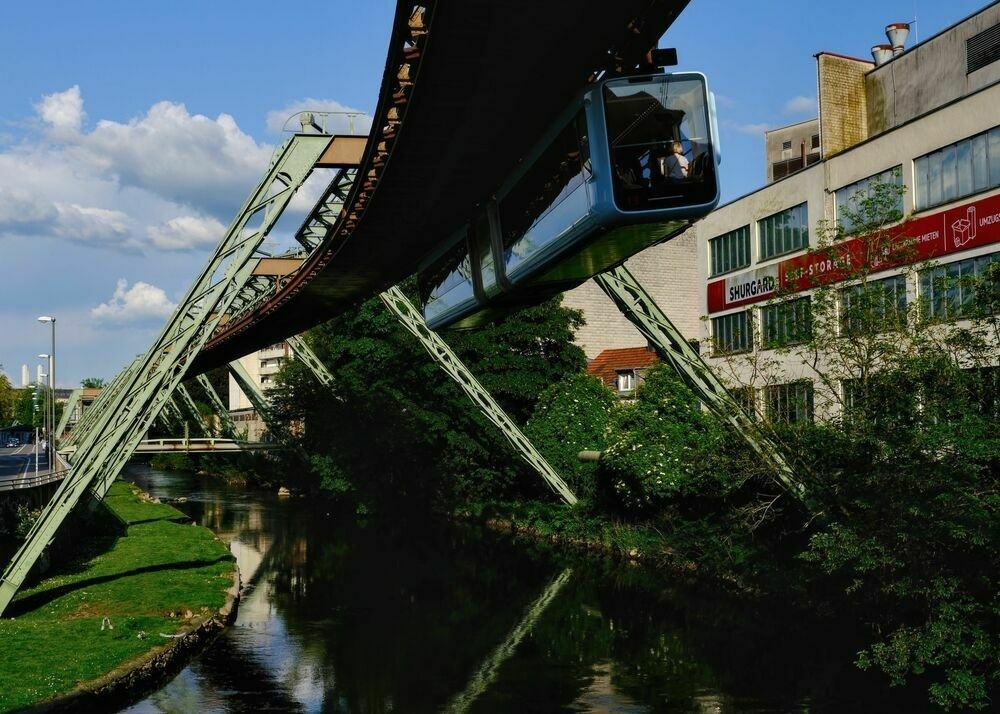 A train approaching overhead, with the angular green metal supporting structure keeping the railway suspended above a river.