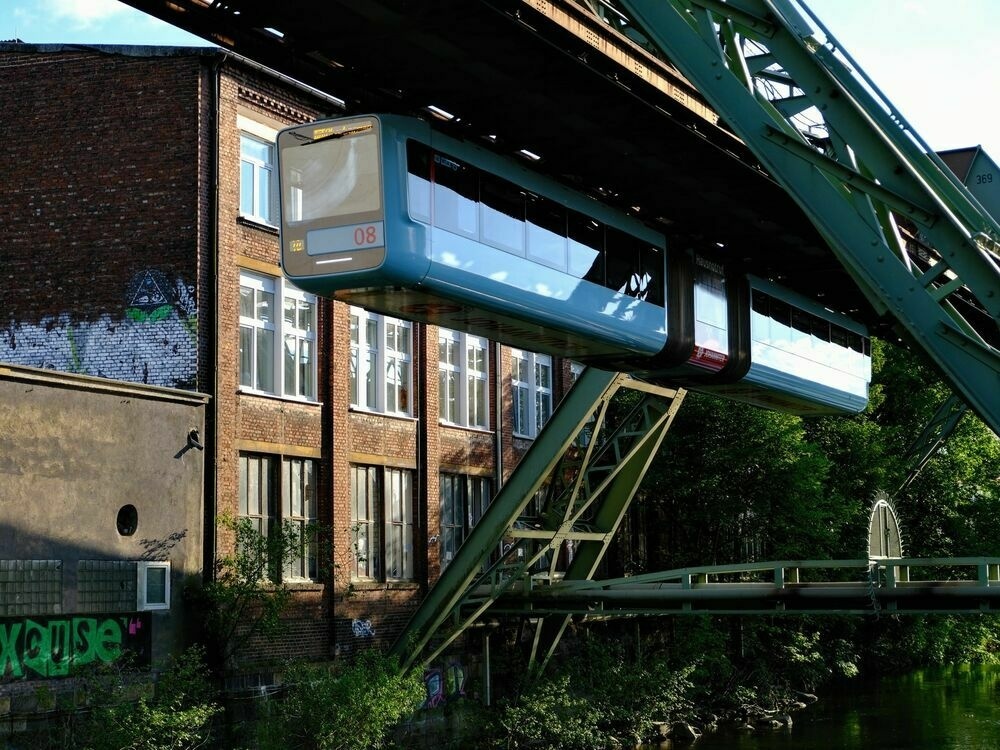 A blue train carriage seen in front of a factory building and above lush green trees and foliage around the river bed.