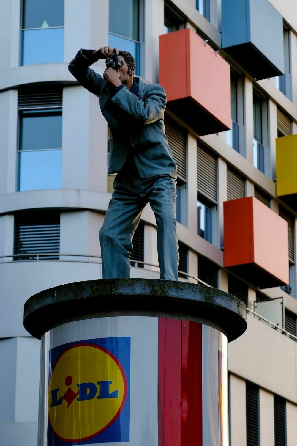 A painted statue of a male figure atop and advertising pillar taking a photograph. Behind him, a colourful building featuring blue, red, and yellow extruding blocks.