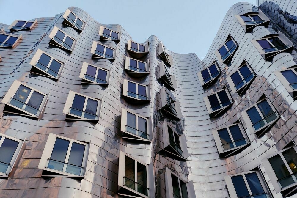 Looking up at a very curvy and reflective building with contrasting angular windows protruding from it.