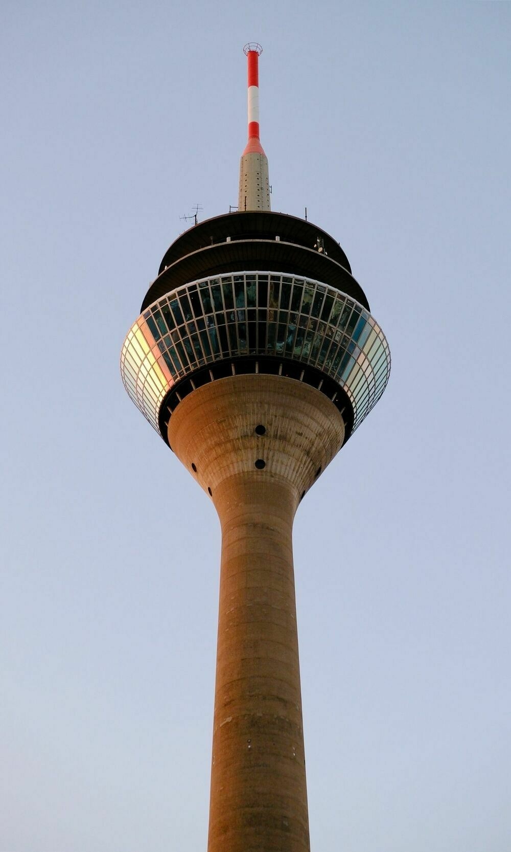 Looking up at a tall and slender concrete communications tower with two glass-frontend floors that spread outwards near the top. A red and white mast sits on the very top of the structure.