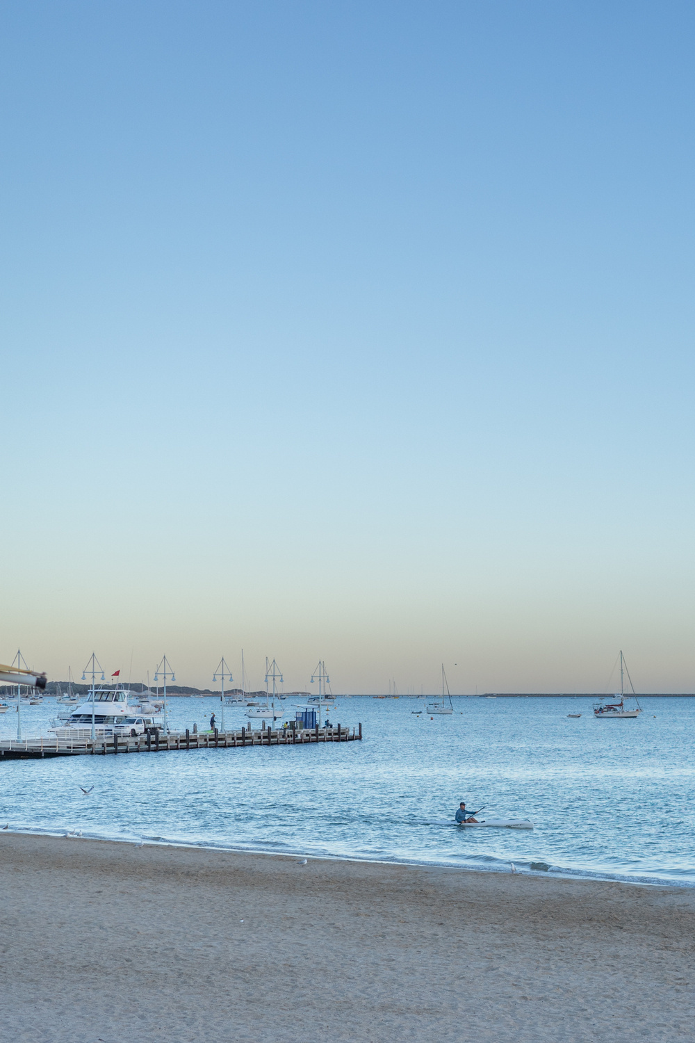 Beach scene with a clear sky at dusk, featuring a person kayaking in the sea, a pier with people, sailboats in the distance, and a boat dock on the left.