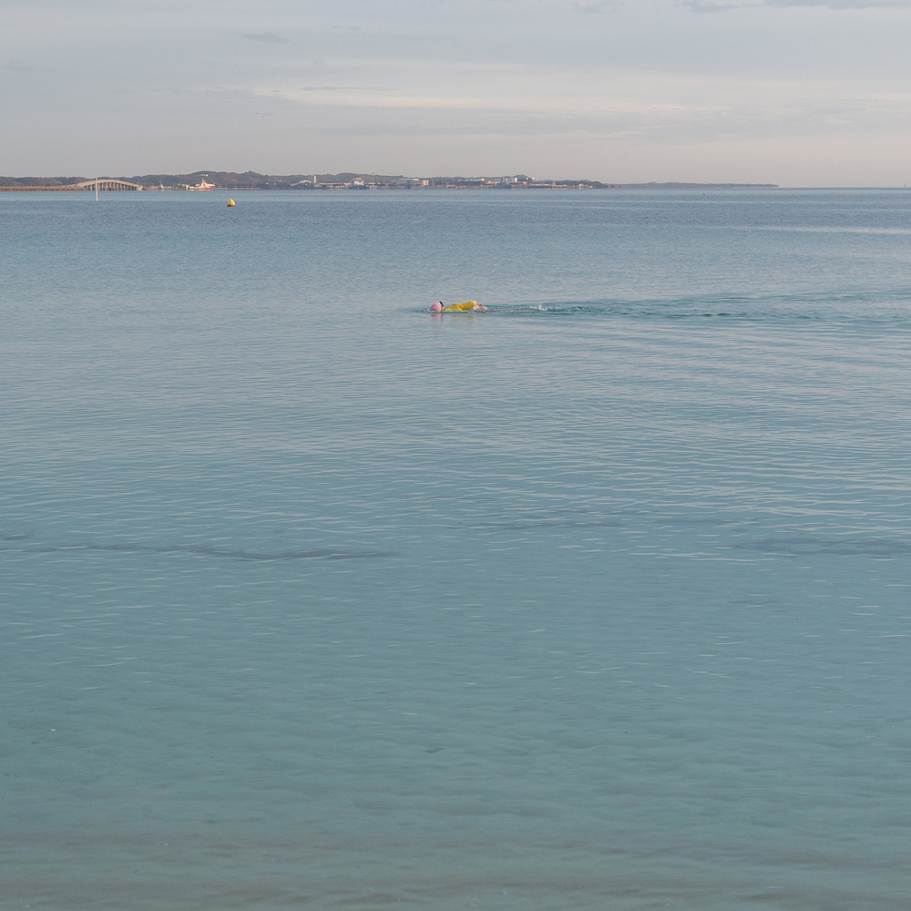Auto-generated description: A person is swimming in a calm blue sea with a buoy nearby, and a bridge connects the distant landmasses under a soft cloudy sky.