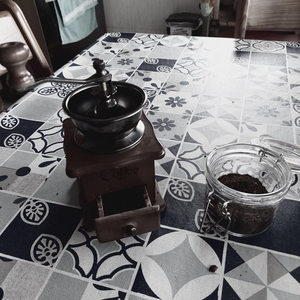 Moinho de café tradicional, de madeira, com o depósito do café moído cheio e aberto. Ao lado, frasco de vidro com café moído. 