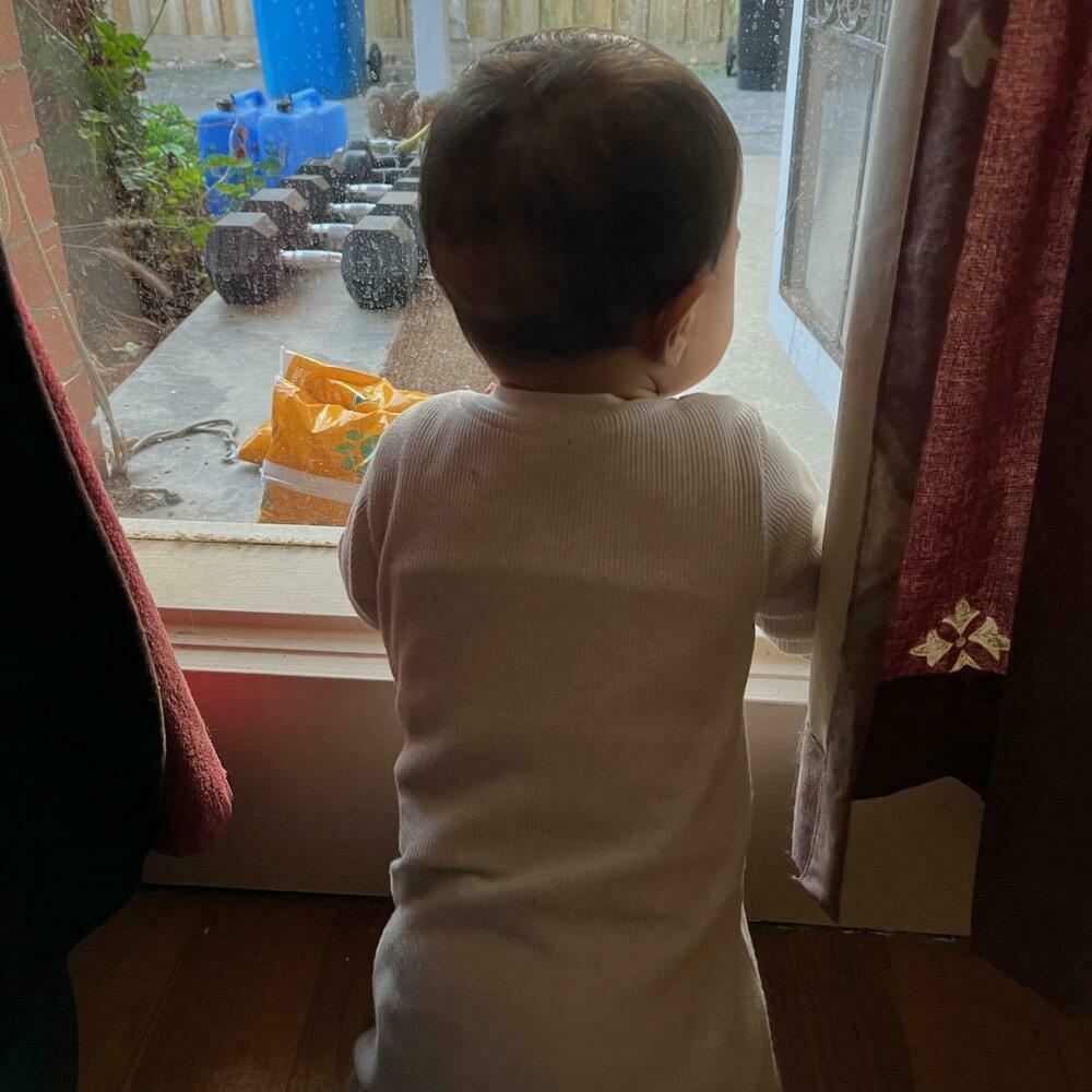 A baby seen from behind kneeling at a window, gazing out wistfully over a cluttered front porch
