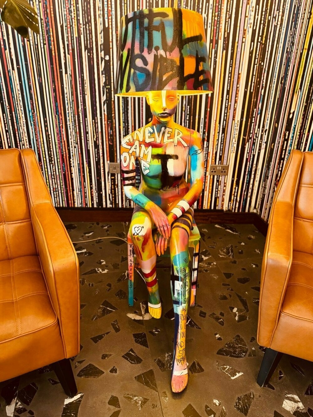 A colorful graffiti-style mannequin sitting between two leather chairs, with a large lampshade over its head, against a wall of vinyl records.