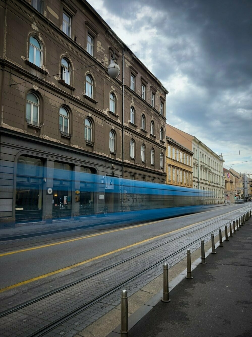 This photo captures a street scene in Zagreb, Croatia. The image shows a slightly worn but charming multi-story building with a faded brown facade, arched windows, and a slightly weathered appearance. The building's architectural style is characteristic of older European cities, with details such as ornamental window frames and a distinctively aged look.

The street in front of the building is lined with tram tracks, and a blue tram is captured in motion, resulting in a blurred effect due to the speed of the tram and the long exposure used in the photo. This blur adds a dynamic element to the otherwise still scene. The tram is moving along a relatively empty street, which is separated from the sidewalk by a series of short metal bollards.

The sky above is cloudy, with heavy, gray clouds suggesting an overcast or stormy day. The overall mood of the image is somewhat moody and quiet, with the tram providing the only sense of movement in an otherwise still urban setting.