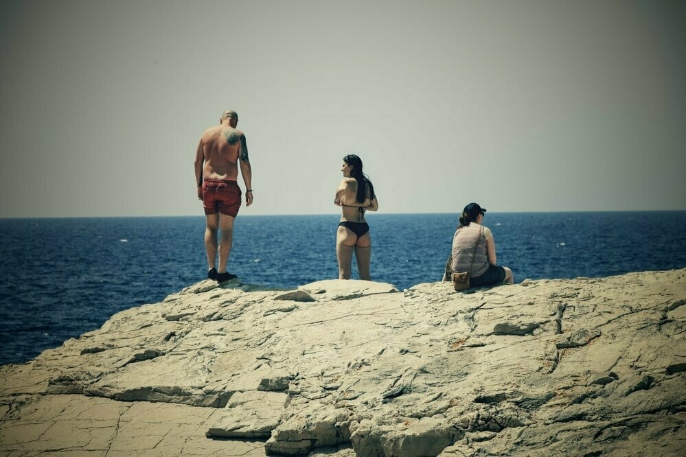 This image captures a scene at the edge of a rocky cliff overlooking the sea. Three people are visible: 

1. On the left, a man with a bald head, wearing red swim trunks, is standing with his back to the camera, looking down at the rocks. He has a tattoo on his upper left arm.
2. In the center, a woman in a black bikini is standing with her arms crossed, facing towards the man and the sea. Her long hair is loose.
3. On the right, another woman is sitting on the rocks, wearing a patterned tank top, shorts, and a black cap. She is facing away from the camera, looking out at the sea.

The sky is clear and the sea is a deep blue, giving the impression of a warm, sunny day. The overall mood of the image is relaxed and contemplative.