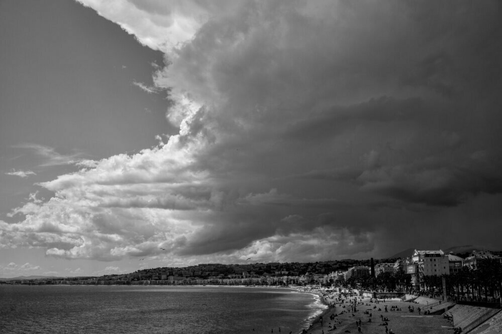 This black and white photograph captures a dramatic coastal scene with a beach, calm ocean, and a cityscape. The sky is the most striking feature, showcasing a stark contrast between dark storm clouds on the right and lighter clouds with some clear sky on the left. The beach, lined with palm trees and lightly populated with people, curves along the shoreline. The city in the middle ground features various buildings, adding to the dynamic atmosphere of the image. The overall composition highlights the contrast between calm and stormy weather.