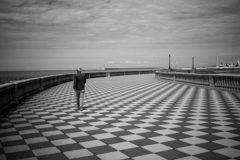 This black and white photograph captures a solitary figure walking along a seaside promenade with a striking checkerboard floor pattern. The person, an older man dressed in a dark jacket and light pants, is seen from behind, walking away from the camera. The promenade curves gently to the right, bordered by a balustrade on the left side, which offers a view of the sea. Several lamp posts line the path, leading towards a distant lighthouse and various structures along the shoreline. The sky is mostly cloudy, with some patches of lighter clouds scattered across. The overall composition evokes a sense of solitude and calmness.