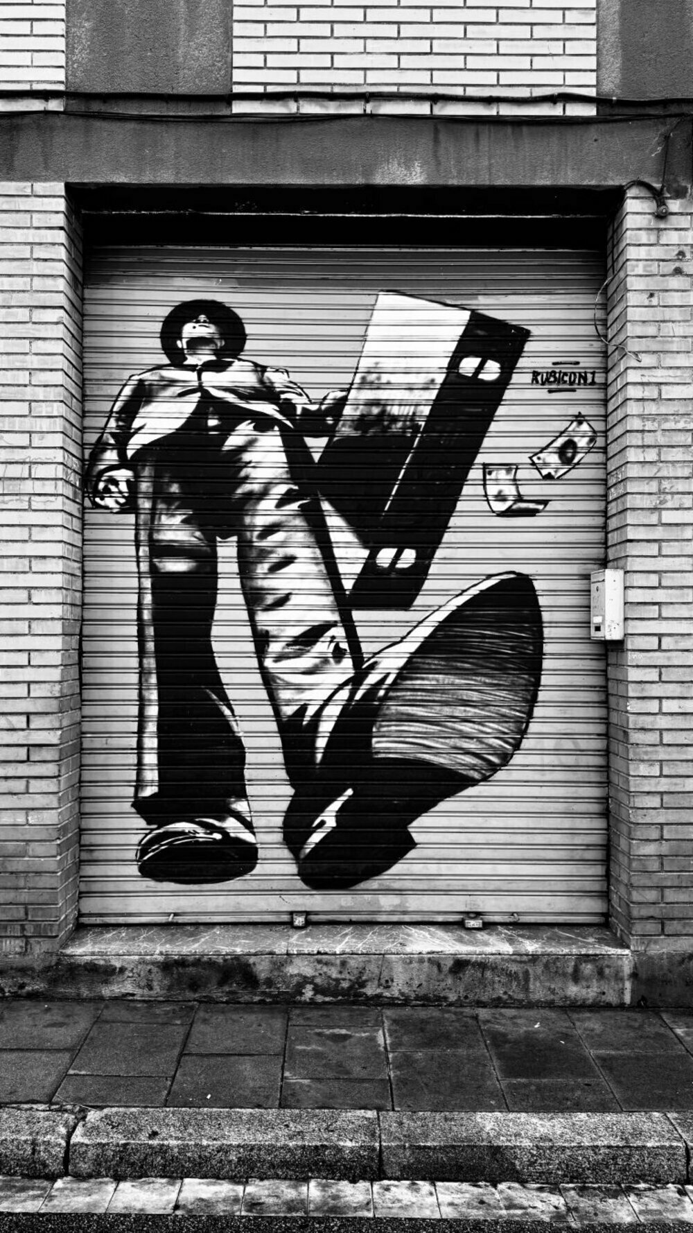 The image depicts a piece of black-and-white street art painted on a metal roll-up door. The mural features a larger-than-life figure of a man in an exaggerated low-angle perspective, making him appear imposing and dominant. The man is wearing a hat and a long coat, reminiscent of a classic film noir or gangster style. He is holding a large, old-fashioned film reel in one hand, and money bills are floating around him, emphasizing a possible theme of wealth or crime.

The door itself is part of a brick building, with horizontal lines from the door's slats adding texture to the artwork. The artist's signature 