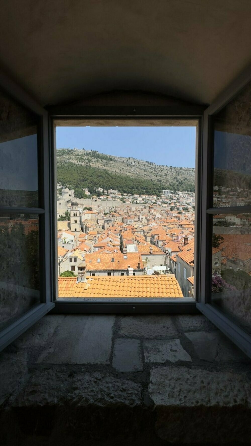 This image shows a scenic view through a window, likely taken from inside an older stone structure. The window frames a picturesque town with numerous closely packed buildings, all featuring distinct red-orange terracotta rooftops. In the distance, you can see a green hillside dotted with trees and small buildings, while the town appears nestled against the base of the hill. The architecture suggests a historic or Mediterranean setting, with stone buildings and narrow streets. The sky is clear and blue, creating a vibrant contrast with the earthy tones of the town and surrounding landscape.