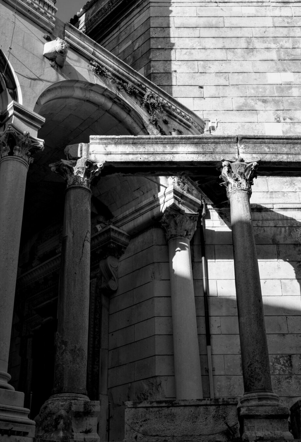 This black-and-white photo depicts part of an ancient stone building with classical architecture. It features large Corinthian columns with ornate capitals, supporting an arch and entablature. The stone blocks show signs of aging, with cracks and weathered textures. Sunlight creates sharp contrasts, casting dramatic shadows that highlight the building's intricate details. The scene conveys a sense of historical grandeur.