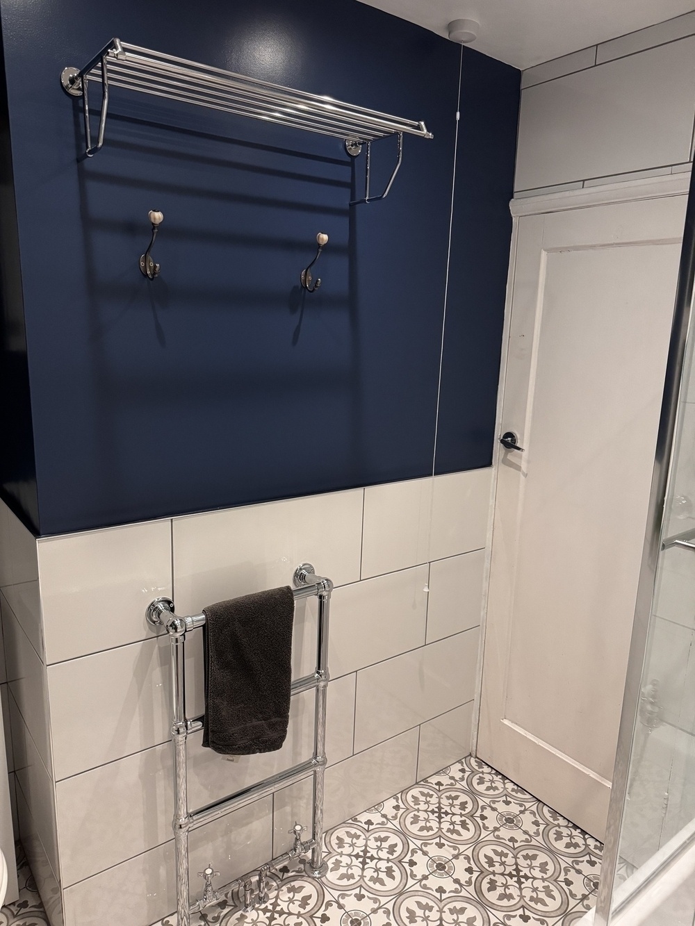 A bathroom features a blue and white tiled wall with a chrome towel rack, hooks, and a shelf, alongside a white door and patterned floor tiles.