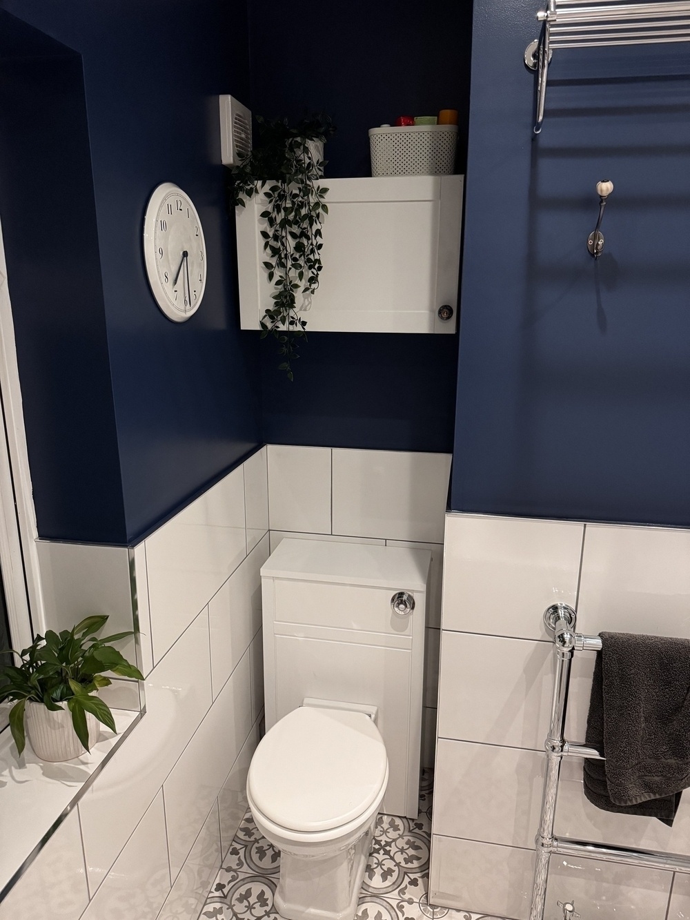 A small bathroom features a white tiled wall, a toilet, a white cabinet with greenery on top, and a dark blue upper wall with a white clock.