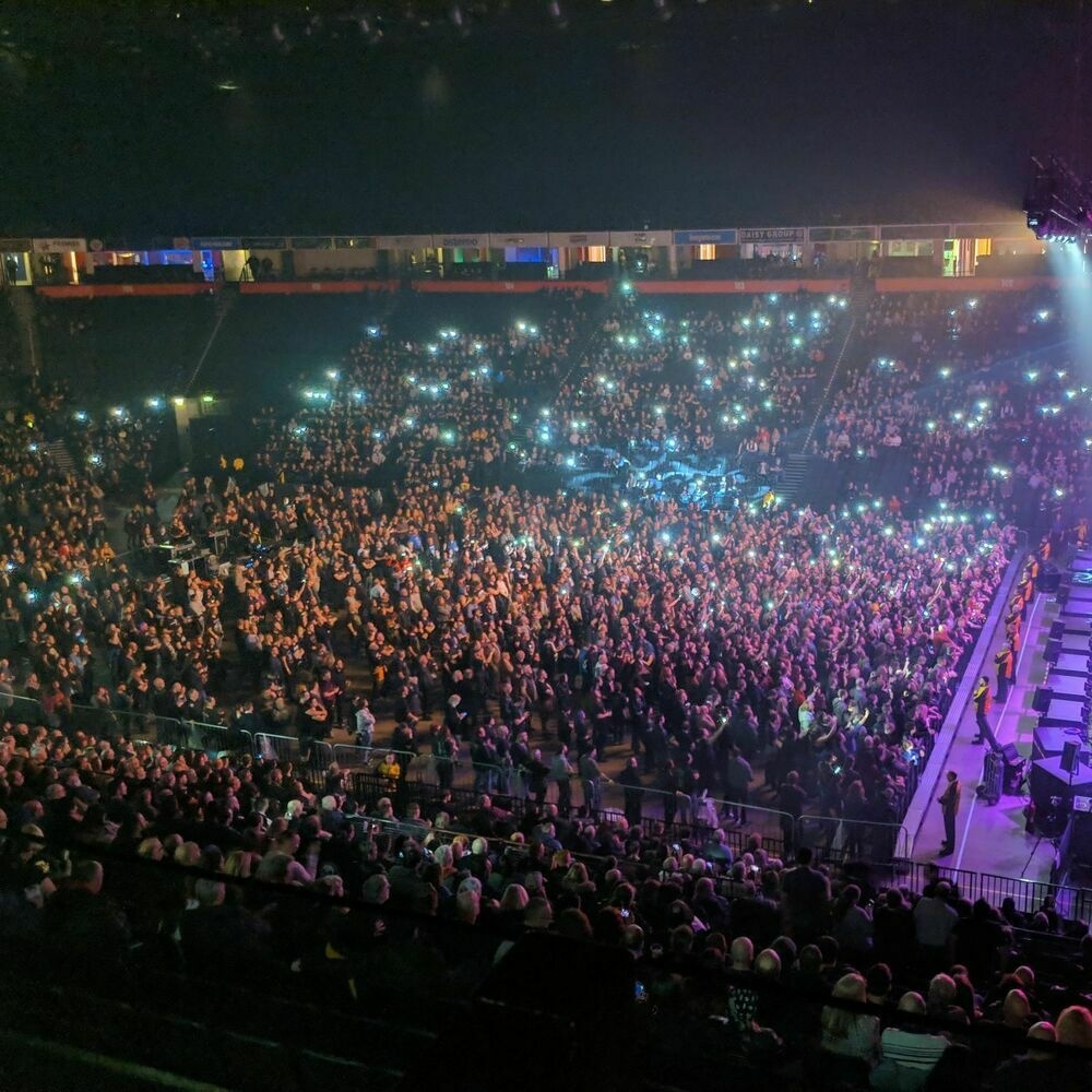 An arena half-full of people attending for a concert, during the supporting band. Google's Night Sight feature is enabled, bringing light into a low-light environment.
