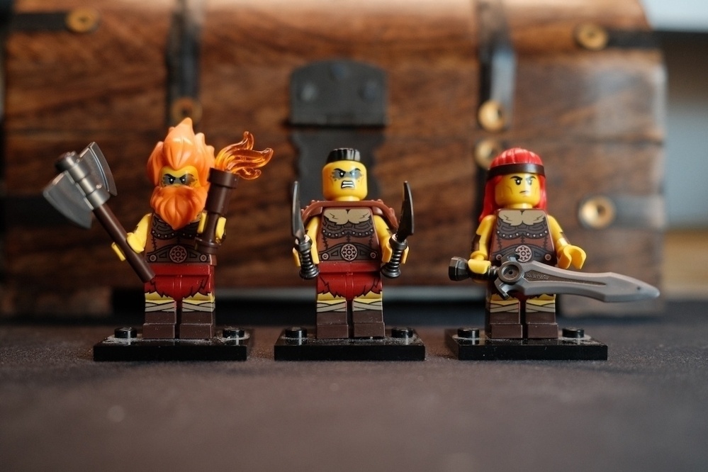 Three Lego warrior minifigures, each equipped with weapons and armor, stand in front of a wooden treasure chest.