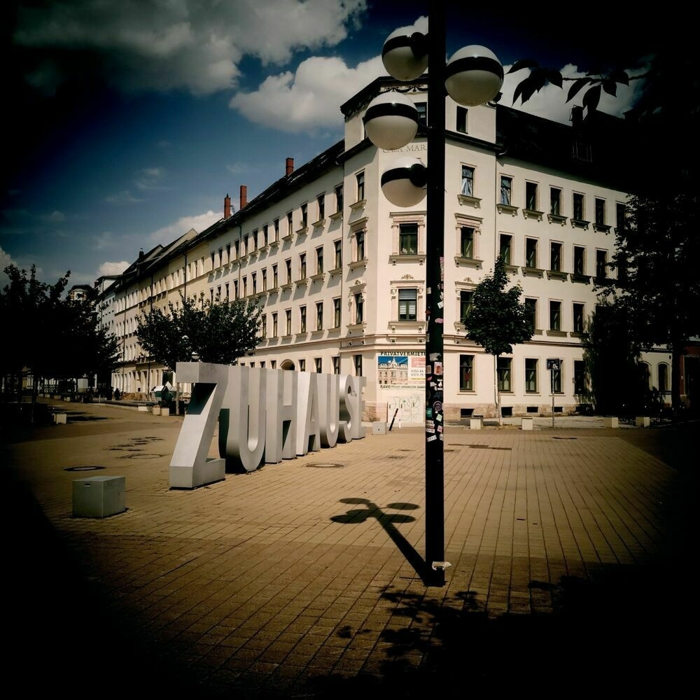 Bright light on a square surrounded by old house facades. There's a lantern in the middle, casting a thin strong shadow, partially on a set of huge letters reading ZUHAUSE which is HOME im German.