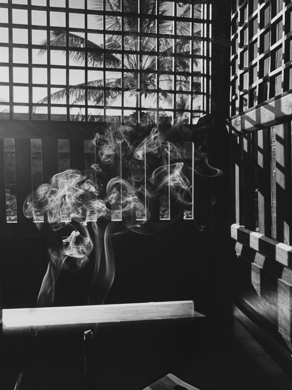 black and white photo of two almost-obscured incense sticks burning wispy trails of smoke appearing light and insubstantial against the dark silhouette of wood carpentry of slats and a square screen casting crossed linear shadows against itself from the steeply-angled and high-contrast light of morning sun, with a coconut tree visible in the background against the sky.
