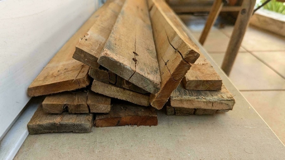 Several pieces of weathered, stacked wooden planks lean against a wall on a tile floor adjacent to a wooden structure.