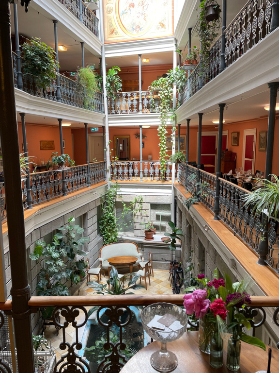 The atrium of the hotel photographed from the first floor. On each of the floors there are growing plants and from the back top part of the image the sun shines into the room. Three floors are visible on the image.