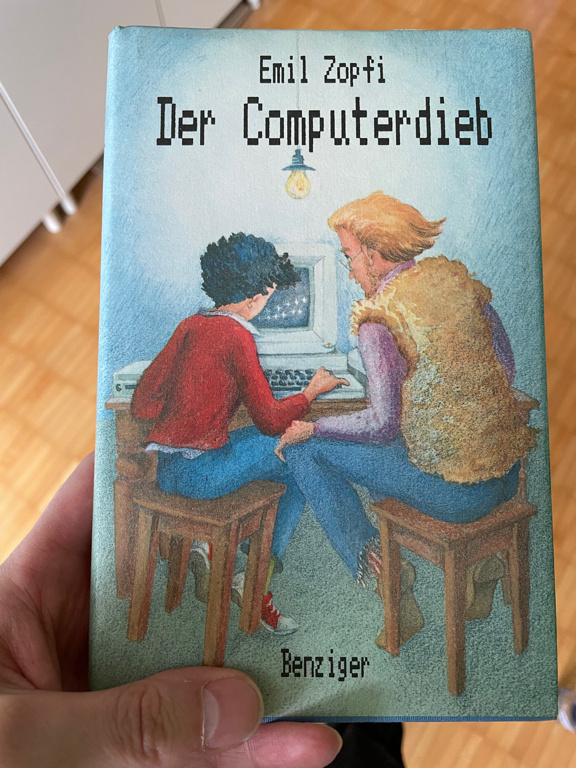 A closeup of book hold in a left hand. It shows two persons sitting on front of a computer. The person on the left is a child with black hair and a read pullover and the person on the right is an adult with blonde hair and a light brown shirt. Both are wearing blue jeans. A the top of the book is the the name of the Author: Emil Zopfi and the book title in German: The Computerdieb (The computer thief).