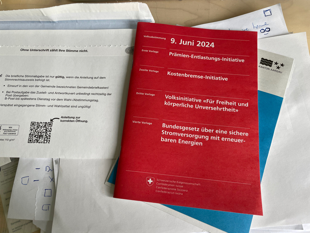 A red booklet on a desk with white text on top. On the left side is the envelop to send it back. The booklet explains the different votes we can vote for on the ninth of June 2024. All text is in German (Präminen-Entlastungs-Initiative, Kostenbremse-Initiative, Volksinitivate Für Freiheit und körperliche Unversehrtheit and Bundesgesetz über eine sicher Stromversorgung mit erneuerbaren Energien).