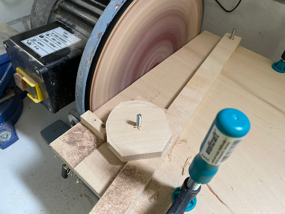 I a closeup of a small disc sander with a circle-making jig clamped in front of it. On top is a raw circle which is int the process of getting sanded round. 