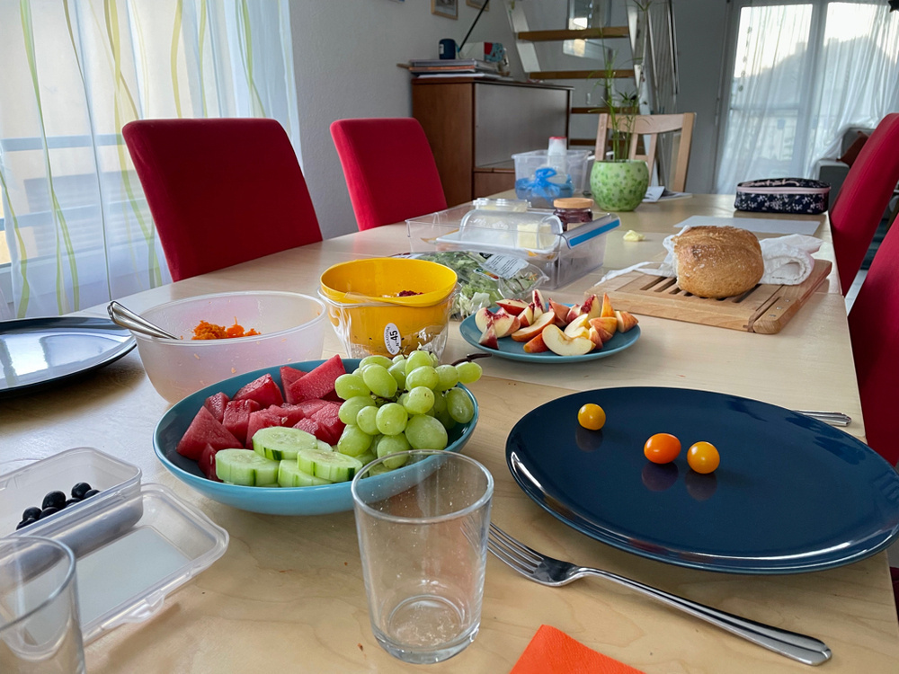 A table spread of different fruits and salads ready to eat. In the front, there are grapes, cucumbers, and watermelon on a plate. Some cherry tomatoes, apples. In the back is a breaks and some green salad. 