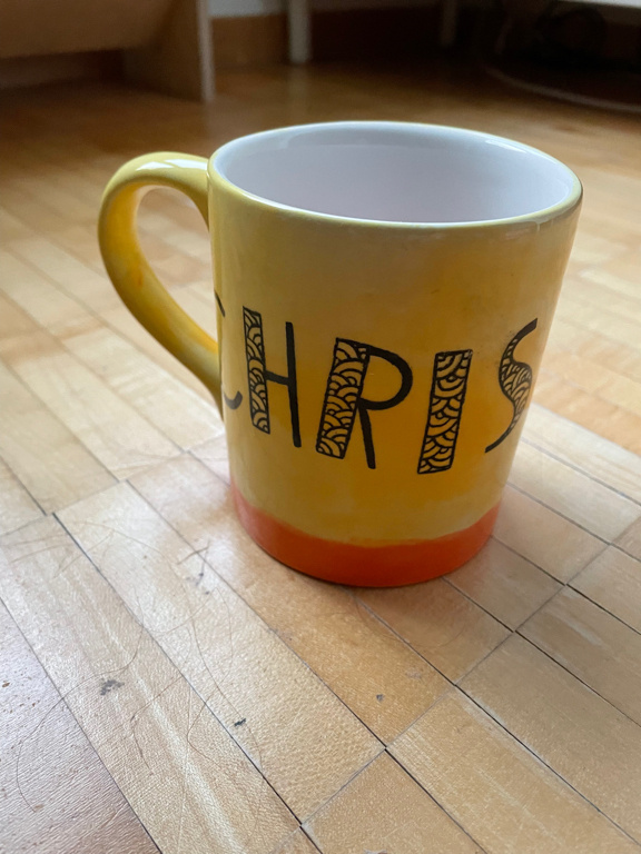 The image shows a yellow ceramic mug with an orange base. The mug has the name "CHRIS" written in large, bold black letters across its side. Each letter has a decorative, patterned design inside. The mug is sitting on a light wooden floor with visible grain and some faint scuff marks. The background includes part of a room with furniture and soft lighting.