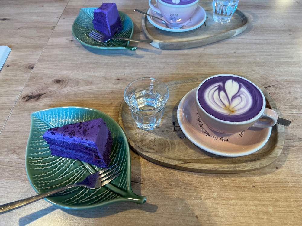 A slice of purple cake is placed on a green, leaf-shaped plate with a fork resting beside it. Next to the plate, a cup of purple-colored latte with white latte art sits on a wooden tray, accompanied by a small glass of water. The items are arranged on a wooden table with visible grain patterns.