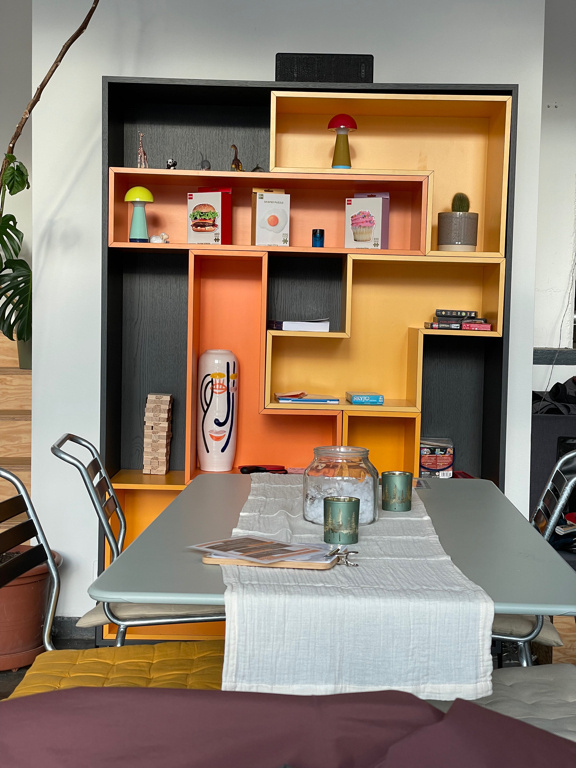 A shelving unit designed to resemble Tetris blocks features colorful compartments in shades of yellow, orange, and black. It displays decorative items such as small lamps, figurines, books, and LEGO sets. A portion of a table with a white table runner and minimal decor is visible in the foreground.
