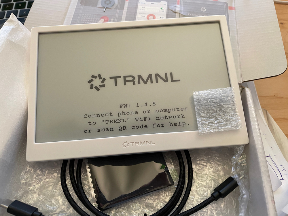 The image shows a TRMNL device with an e-ink display, still partially wrapped in protective packaging. The setting suggests an unboxing scene, possibly in preparation for installation or first-time use. The screen displays setup instructions, indicating firmware version 1.4.5 and directing the user to connect to the "TRMNL" WiFi network or scan a QR code for help. The device has a white frame with the TRMNL logo printed below the screen. A piece of bubble wrap partially covers the lower right corner of the screen. Inside the box, there is a coiled black braided USB-C cable. The box itself has a foam and bubble wrap lining.