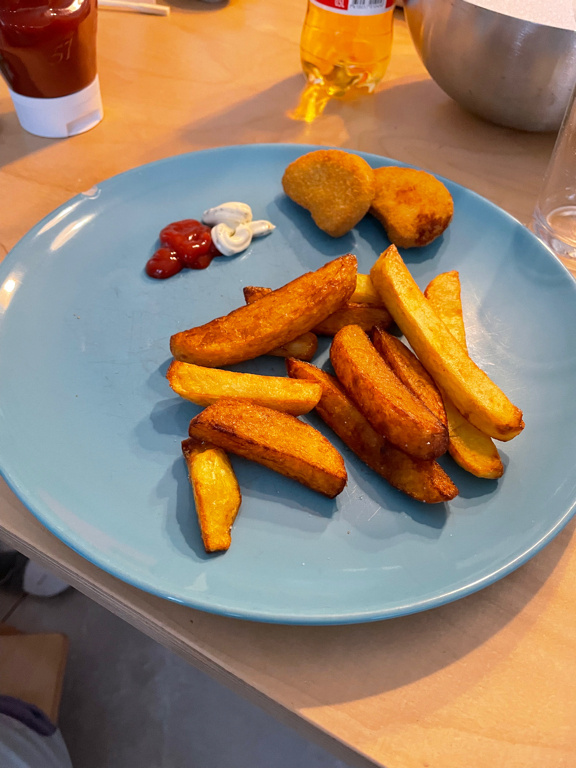 A blue plate with fried potato wedges and chicken nuggets arranged on it. There are small servings of ketchup and mayonnaise on the plate. The background shows a wooden table with a bottle of soda and a metal bowl partially visible.