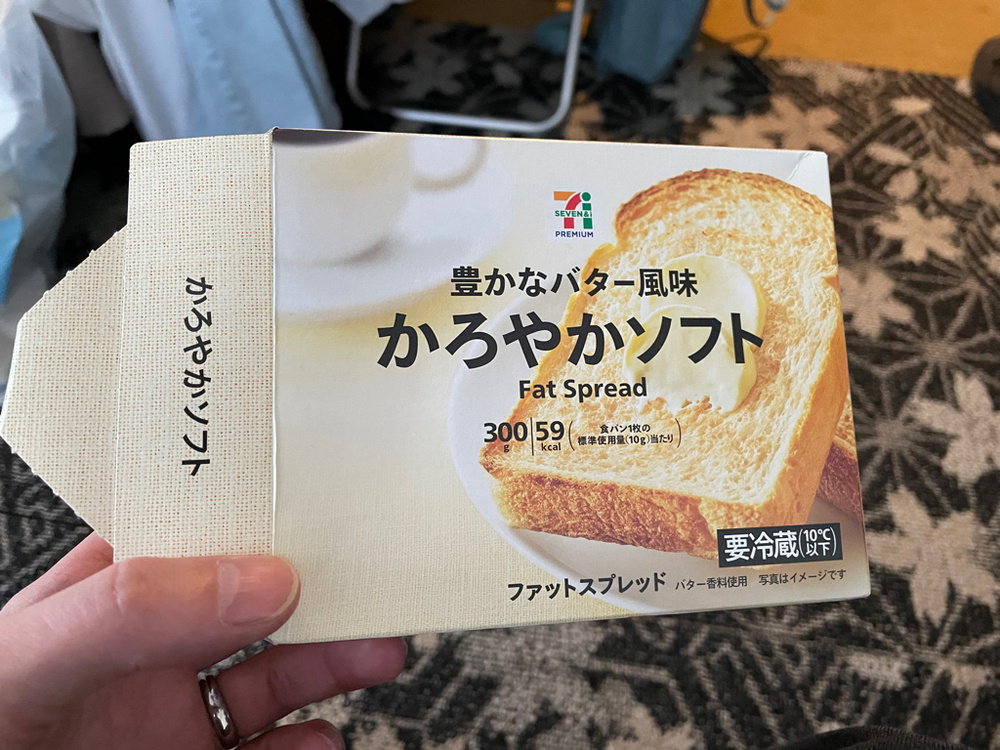 A person holding a package of 7-Eleven Premium fat spread. The packaging features an image of a slice of bread with a dollop of spread. The text includes Japanese characters and states "Fat Spread" along with nutritional information indicating 59 kcal per serving. The design suggests a butter-flavored product.