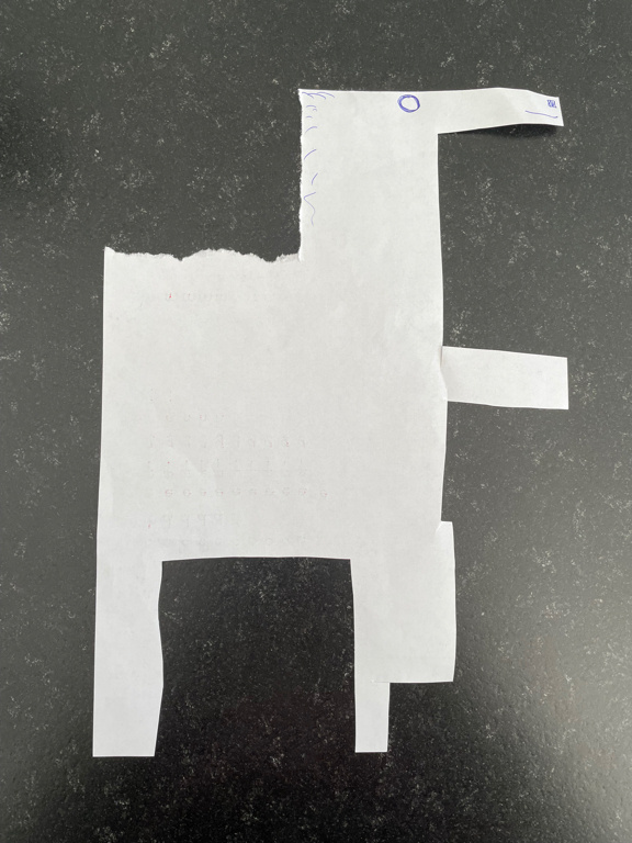 A piece of white paper has been cut into the shape of an abstract animal resembling a horse or a llama. It features a long neck, four legs, and a rectangular body. Simple facial details are drawn on it in blue pen, such as an eye and a few lines representing hair. The paper lays flat on a dark, textured surface.